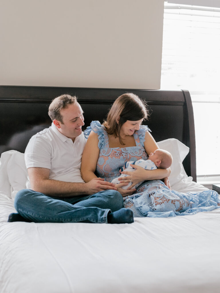 Husband and wife sit cozily on their bed gazing at their newborn baby in the mother's arms