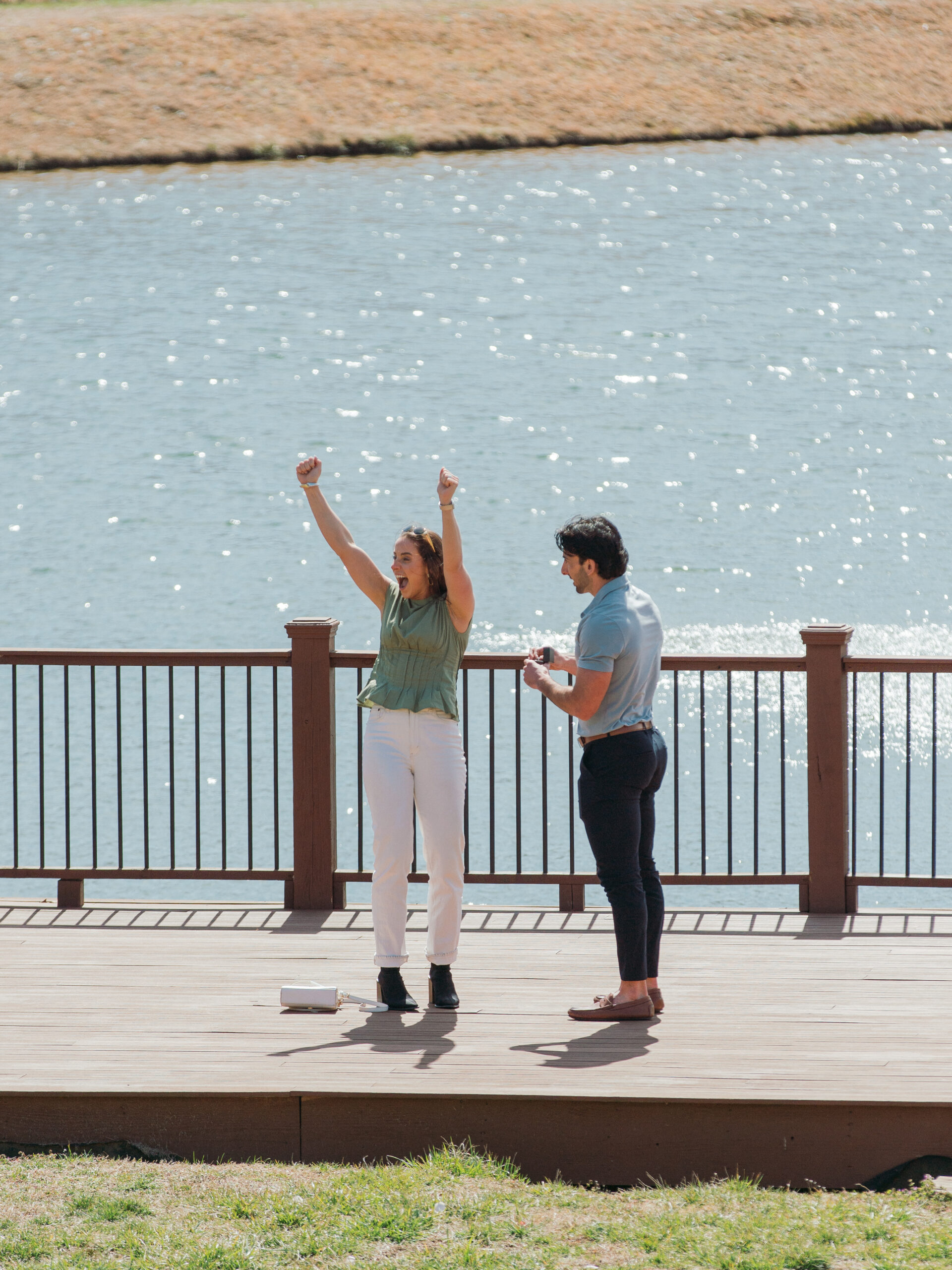 A joyful moment as a woman raises her arms in excitement after a surprise proposal by a lake. Her fiancé, holding the ring box, smiles as he watches her reaction. The scene is set on a wooden deck at Montaluce Winery with a serene water backdrop, capturing the pure happiness of their engagement.